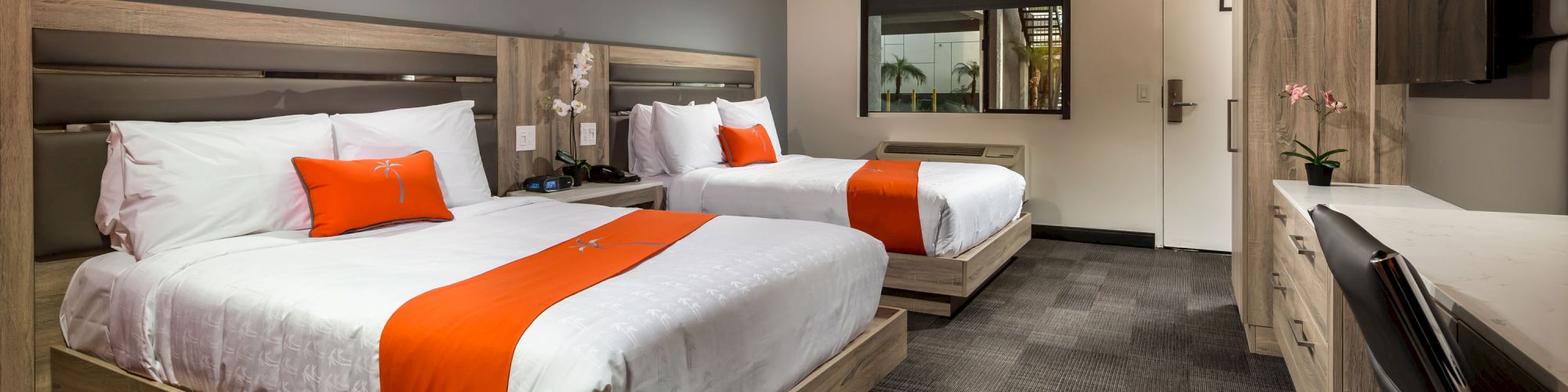 This image shows a modern hotel room with two double beds, white linens, and orange accents. There is a TV, desk, and contemporary decor.