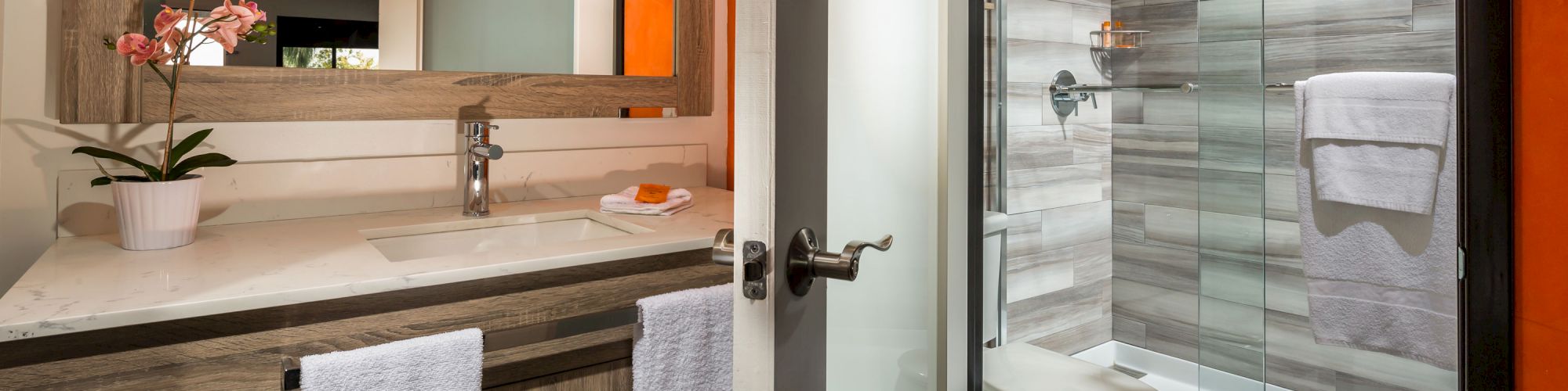 The image shows a modern bathroom with a glass shower, a toilet, a vanity with a sink, a large mirror, white towels, and a potted plant on the counter.