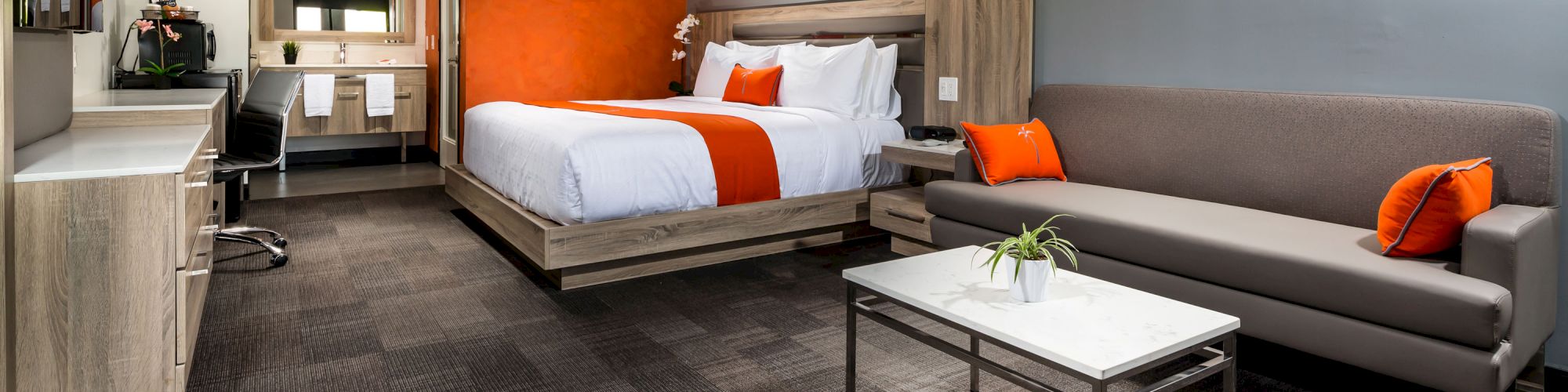 A modern hotel room with a bed, sofa, desk, coffee maker, and TV, featuring grey and orange decor, and an adjoining bathroom visible.