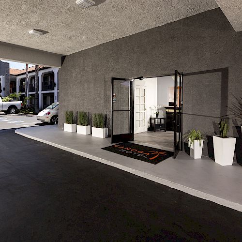 The image shows the entrance to a building with glass doors, a black welcome mat, and several white planters with plants. A parking area is outside.