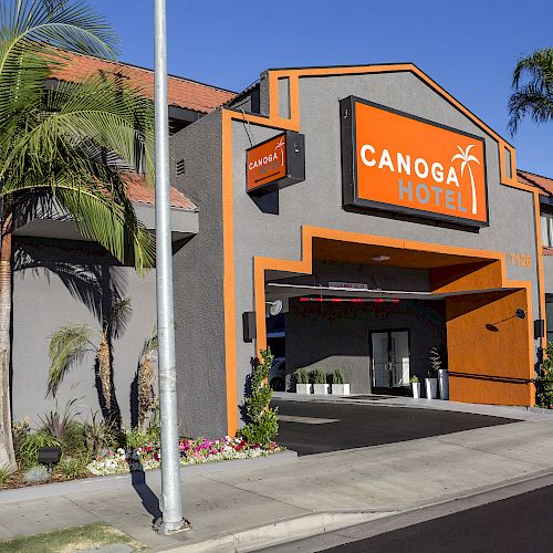 A hotel named Canoga Hotel, with gray and orange exterior, palm trees, a driveway, and a clear blue sky.