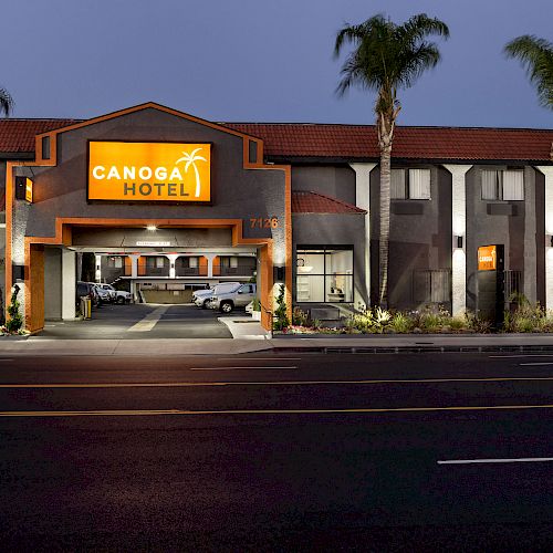 The image shows the exterior of Canoga Hotel at night, featuring a lit-up sign, parked cars underneath, palm trees, and a well-maintained facade.