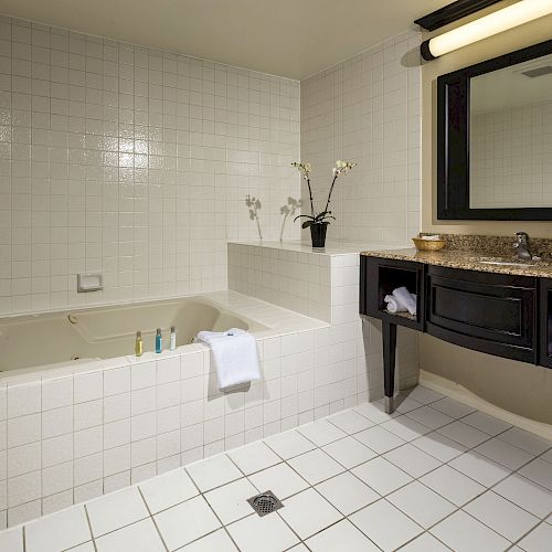 A modern bathroom with a large bathtub, a granite-countertop vanity, a framed mirror, and white tiled walls and floor, ending the sentence.