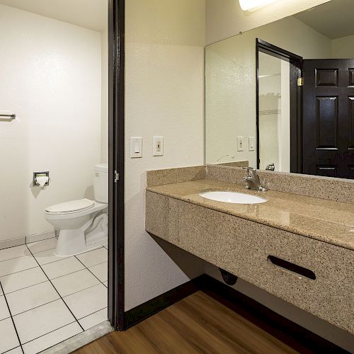 A bathroom with a granite countertop, sink, and mirror, adjacent to a toilet and towel rack, separated by a partial wall and door.