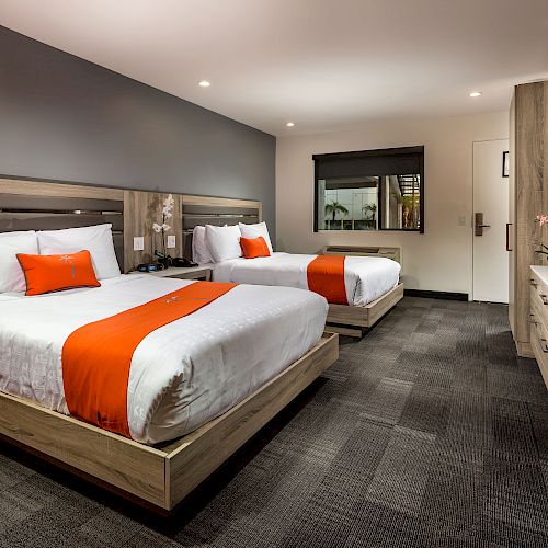 A modern hotel room with two double beds, an orange accent blanket and pillows, a desk, a TV, and a large window.