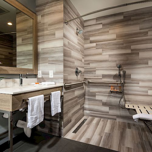 The image shows a modern bathroom with a large mirror, sink, towels, and a walk-in shower with a wooden seat and grab bars, featuring wood-patterned tiles.