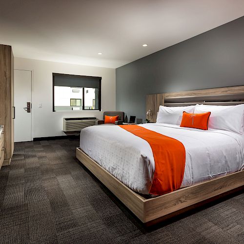 The image shows a modern hotel room with a large bed, a desk, chairs, a wall-mounted TV, and an orange accent blanket and pillow.