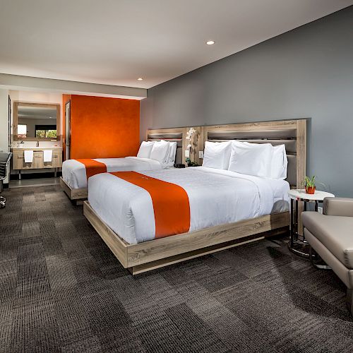 A modern hotel room features two queen beds with orange accents, a desk, a chair, a small seating area, and a coffee maker.