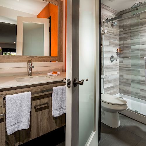 A modern bathroom features a sink with a mirror, towels, a glass-enclosed shower, and a flower vase on the counter, along with a door.