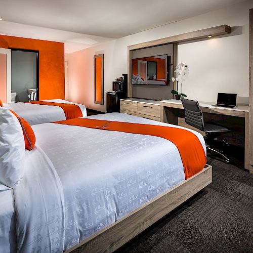 A hotel room with two beds, orange accents, a desk area with a chair, a large mirror, a TV, a wardrobe, and modern decor.