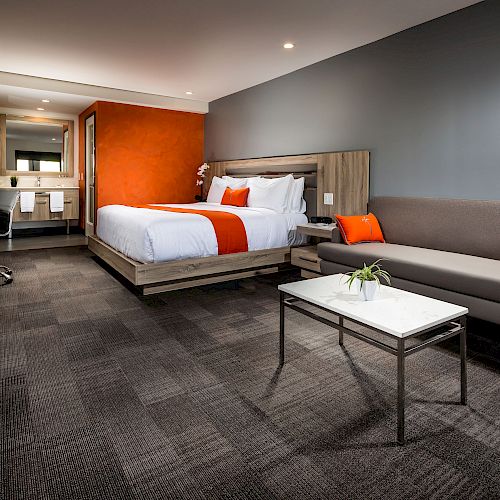 The image shows a modern hotel room with a double bed, a couch, a coffee table, and a desk area with amenities. The colors are grey and orange.