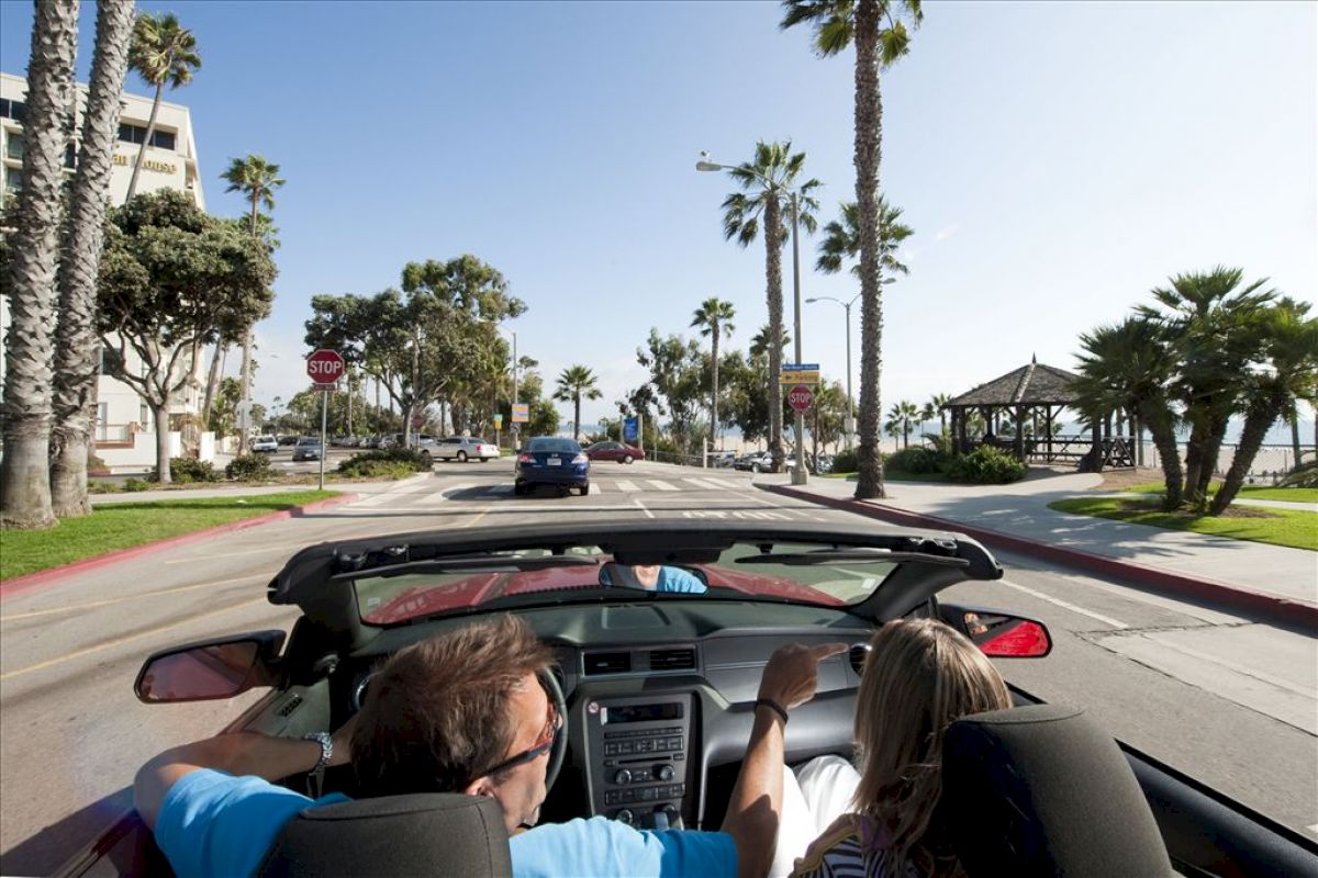 Two people are driving in a convertible on a palm-lined street near the beach, with clear skies and light traffic around them.