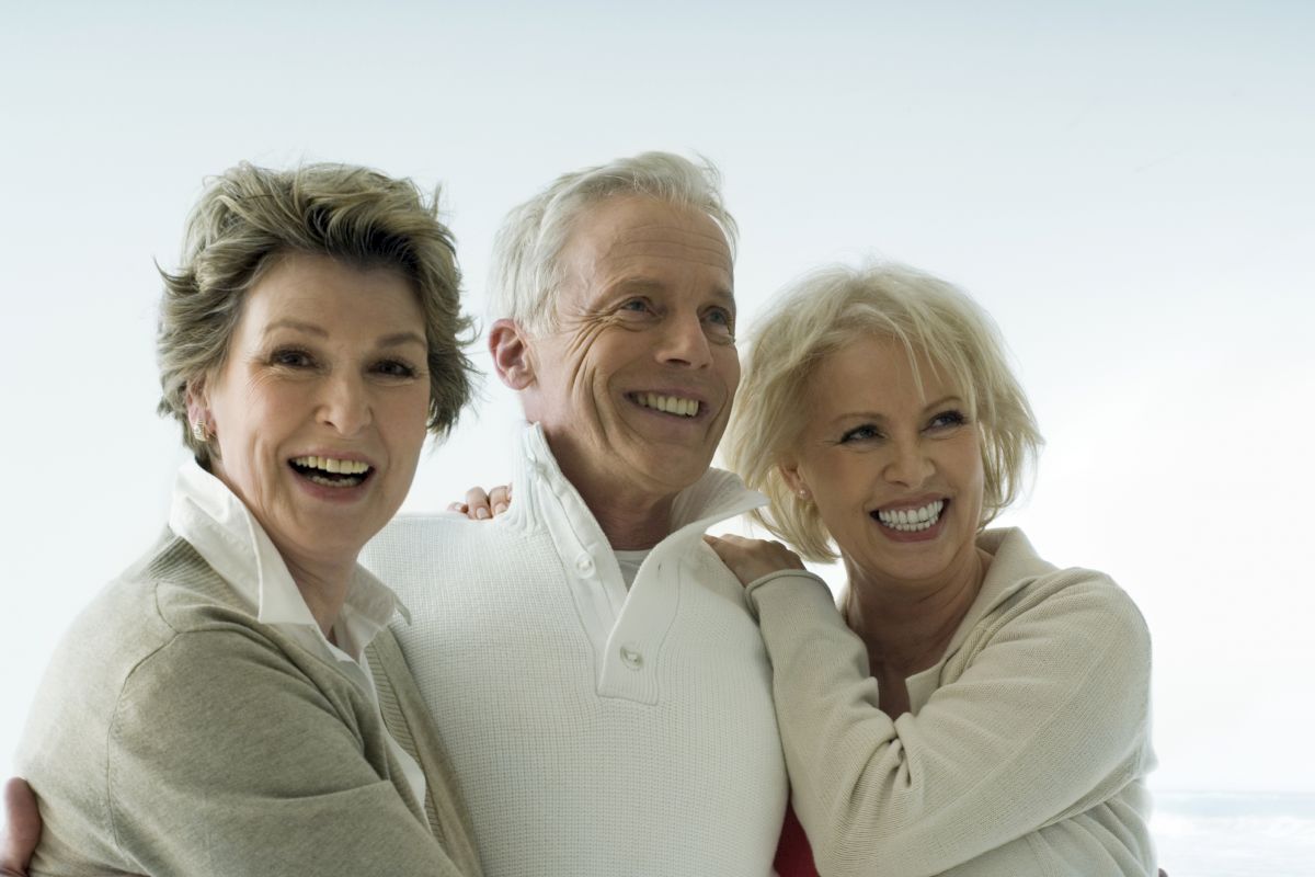 Three older adults, two women and one man, are standing closely together and smiling, appearing happy and joyful against a light background.