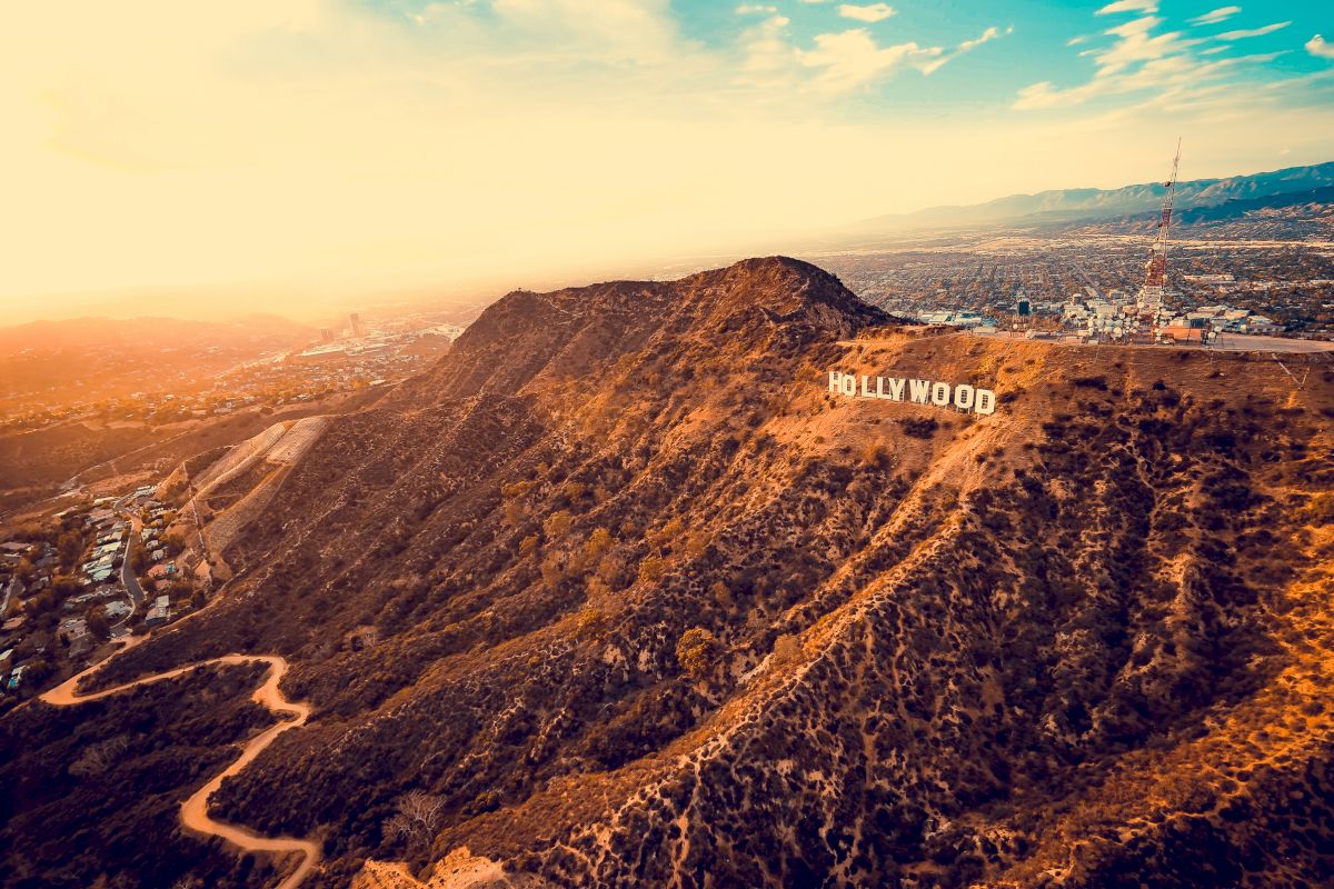 This image captures an aerial view of the iconic Hollywood sign on the hills overlooking Los Angeles at sunset, with the city extending in the background.