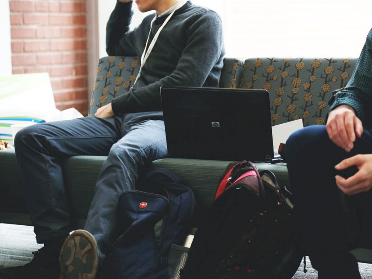 Two individuals are seated on a couch with laptops and backpacks, engaged in conversation or task, indoors with natural lighting.