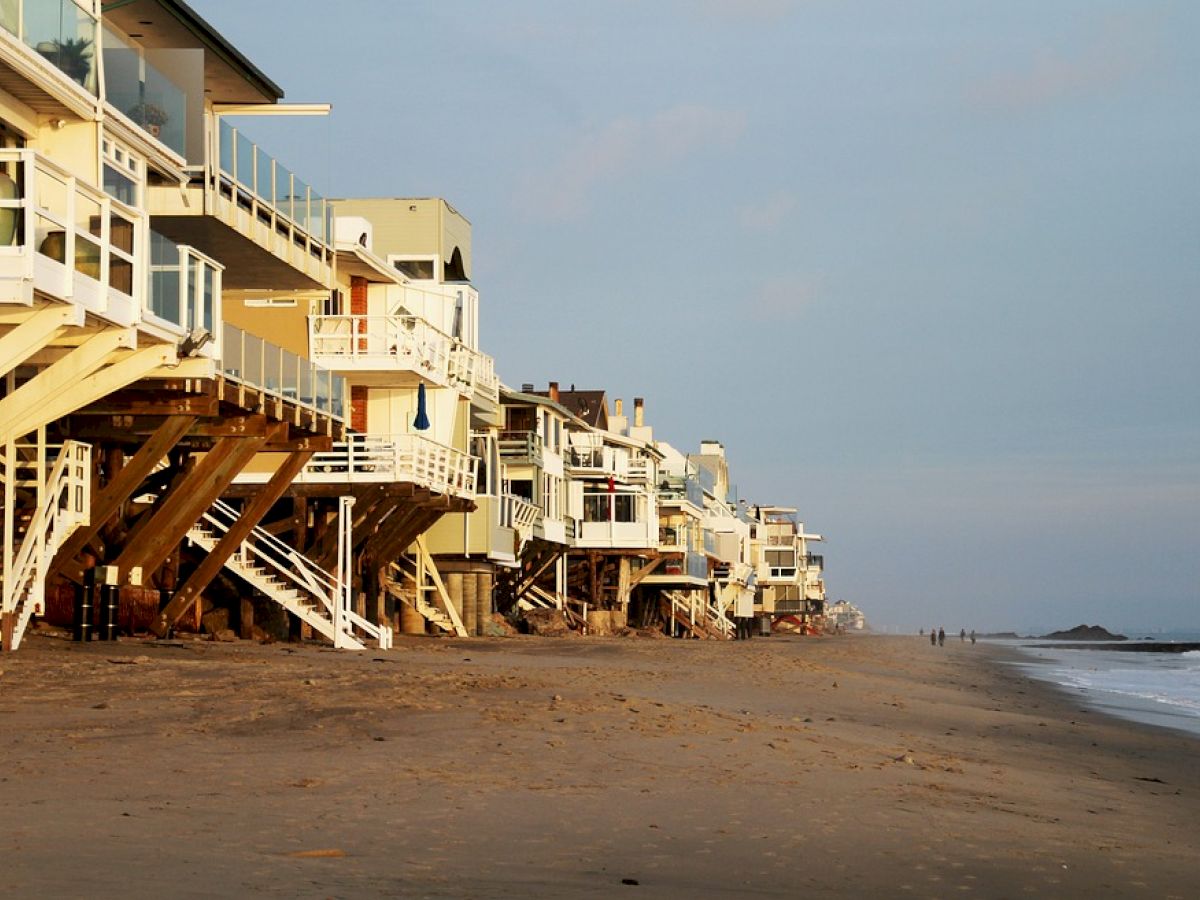 This image shows a row of modern, elevated beachfront houses along a sandy beach, with the ocean visible on the right-hand side.