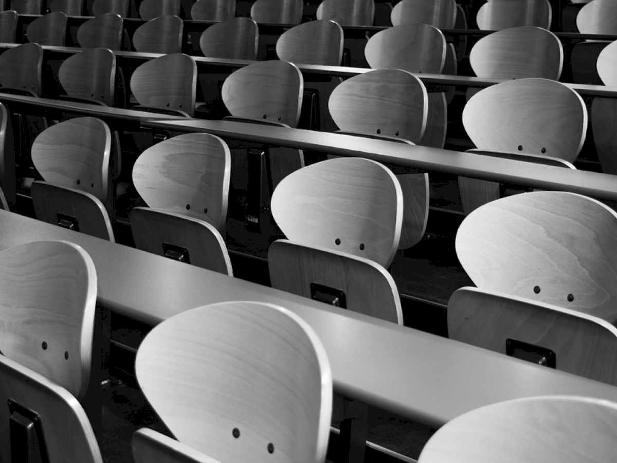 This image shows several rows of empty wooden chairs and long desks in what appears to be a lecture hall or classroom.