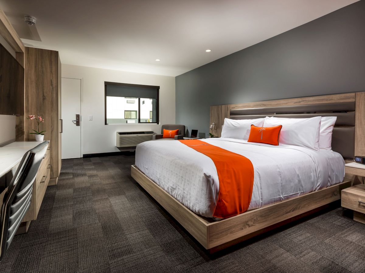 A modern hotel room with a large bed, orange accents, a desk with a chair, a flat-screen TV, and a window with a view of the outdoors.