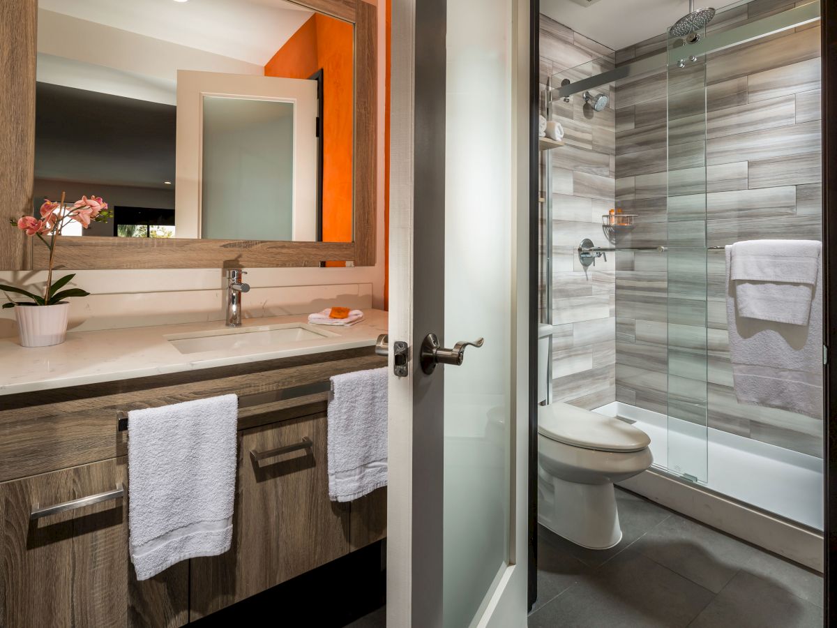 The image shows a modern bathroom with a sink, mirror, potted plant, towels on the rack, a glass-enclosed shower, and white tiled walls.