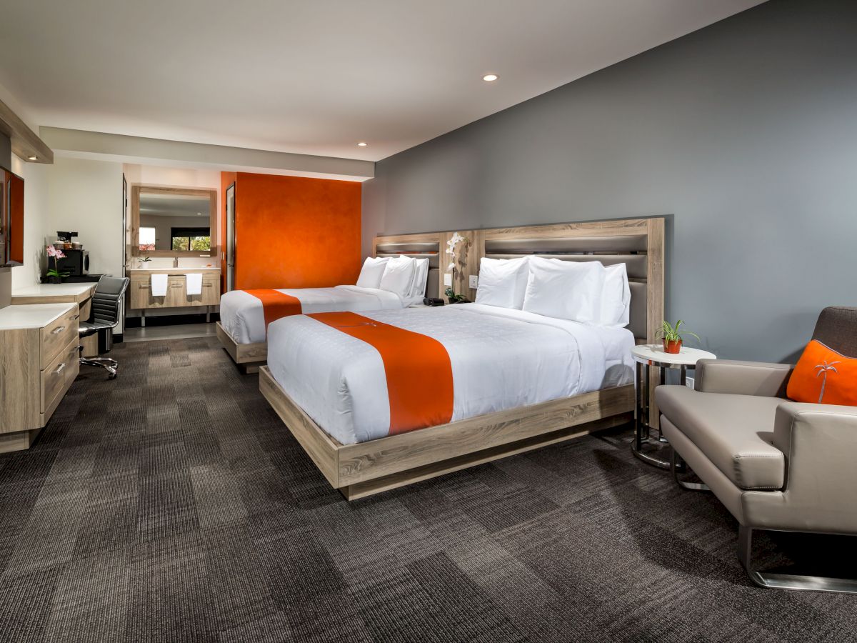 A modern hotel room features two double beds with orange accents, a desk, an armchair, and a bathroom in the background.