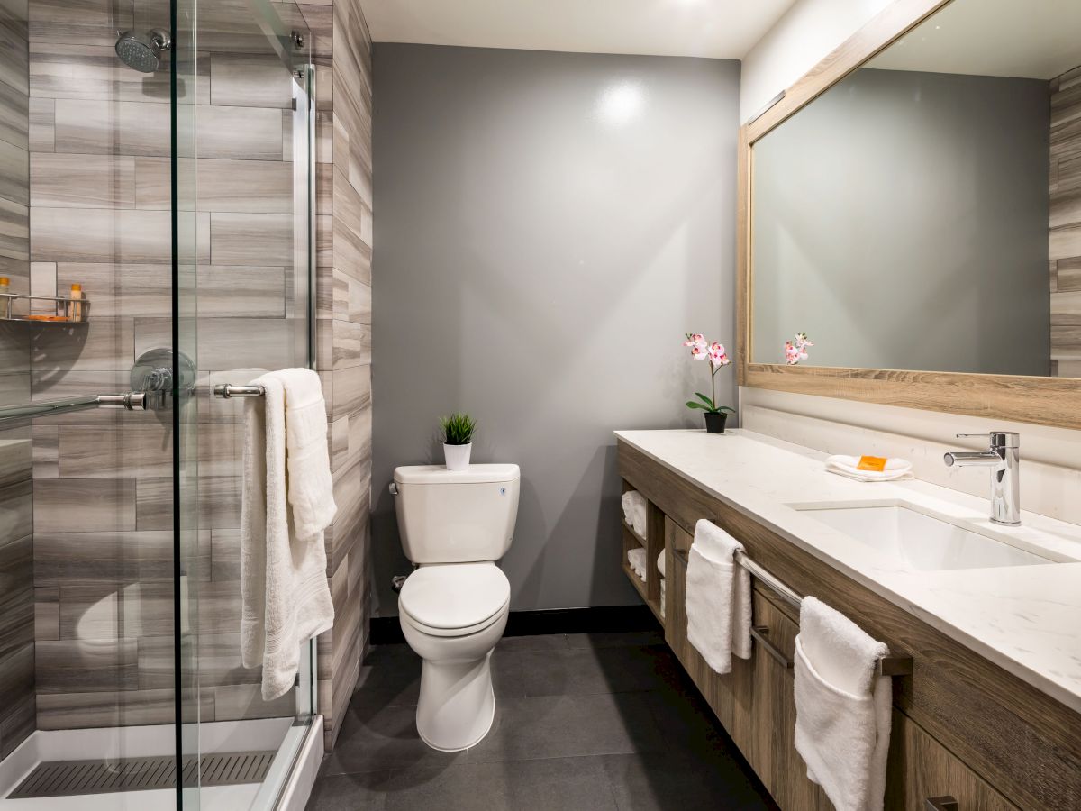 A modern bathroom with a glass shower, toilet, wooden vanity, large mirror, and grey-tiled wall. It has plants and towels as decor.