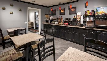 A small room with tables, chairs, wall decor, and a counter with coffee machines, a juice dispenser, and other appliances.