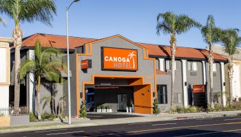 This image shows the exterior of the Canoga Hotel, with a modern design, palm trees, and a clear blue sky in the background.