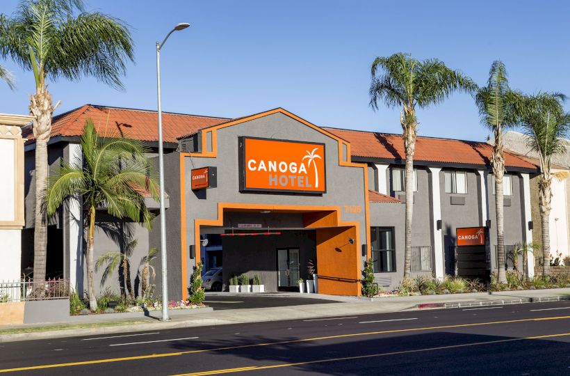 This image shows the exterior of the Canoga Hotel, with a modern design, palm trees, and a clear blue sky in the background.