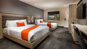 The image shows a modern hotel room with two beds with white linens and orange accents, a TV, a desk, and neutral décor ending the sentence.