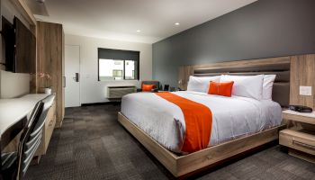 A modern hotel room with a king-sized bed, two orange pillows, and an orange runner, featuring a desk, TV, and air conditioning unit under the window.