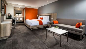 The image shows a modern hotel room with a bed, sofa, coffee table, desk, and amenities. The decor includes orange accents and a sleek design.