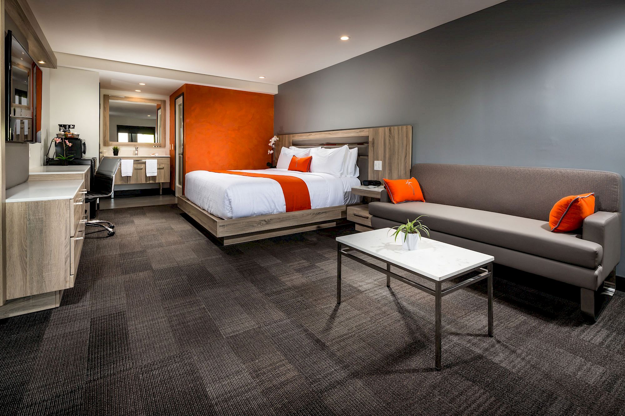 A modern hotel room with a bed, couch, coffee table, desk, and bathroom. The room features a gray and orange color scheme, with clean, minimalist decor.