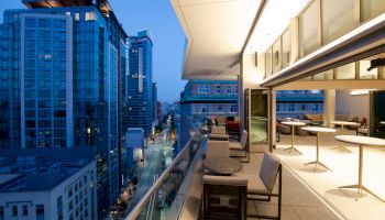 The image shows a modern cityscape with tall buildings, viewed from a balcony with seating and tables. The sky is clear at dusk.