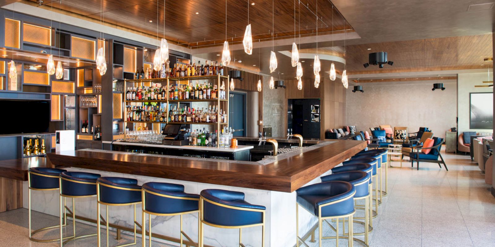The image shows a modern bar with a variety of liquor bottles on shelves, stylish bar stools, hanging lights, and a seating area in the background.