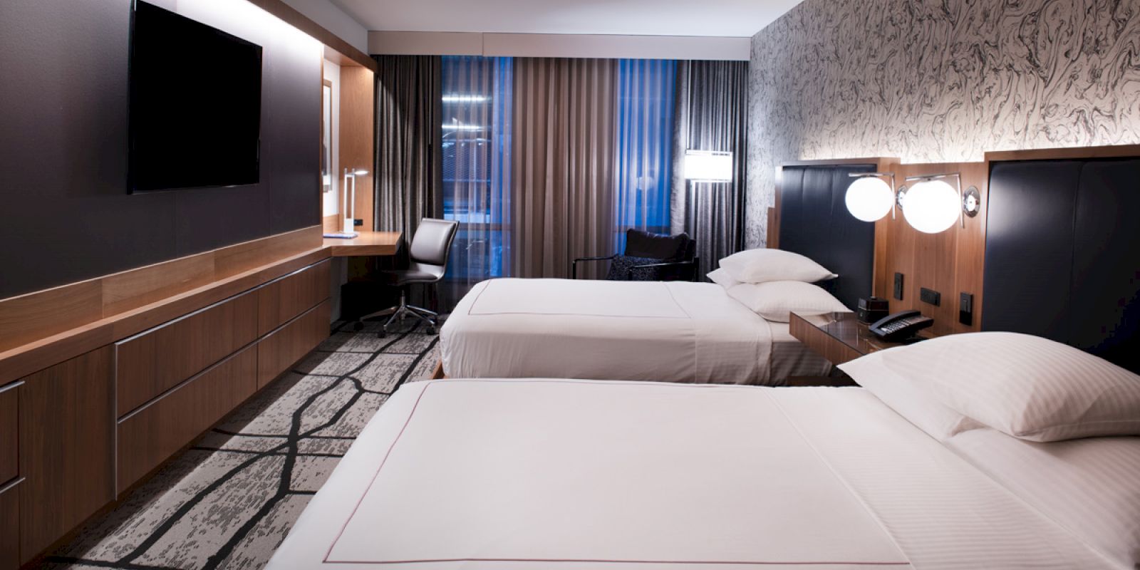The image shows a modern hotel room with two double beds, a desk with a chair, a flat-screen TV on the wall, and stylish decor.