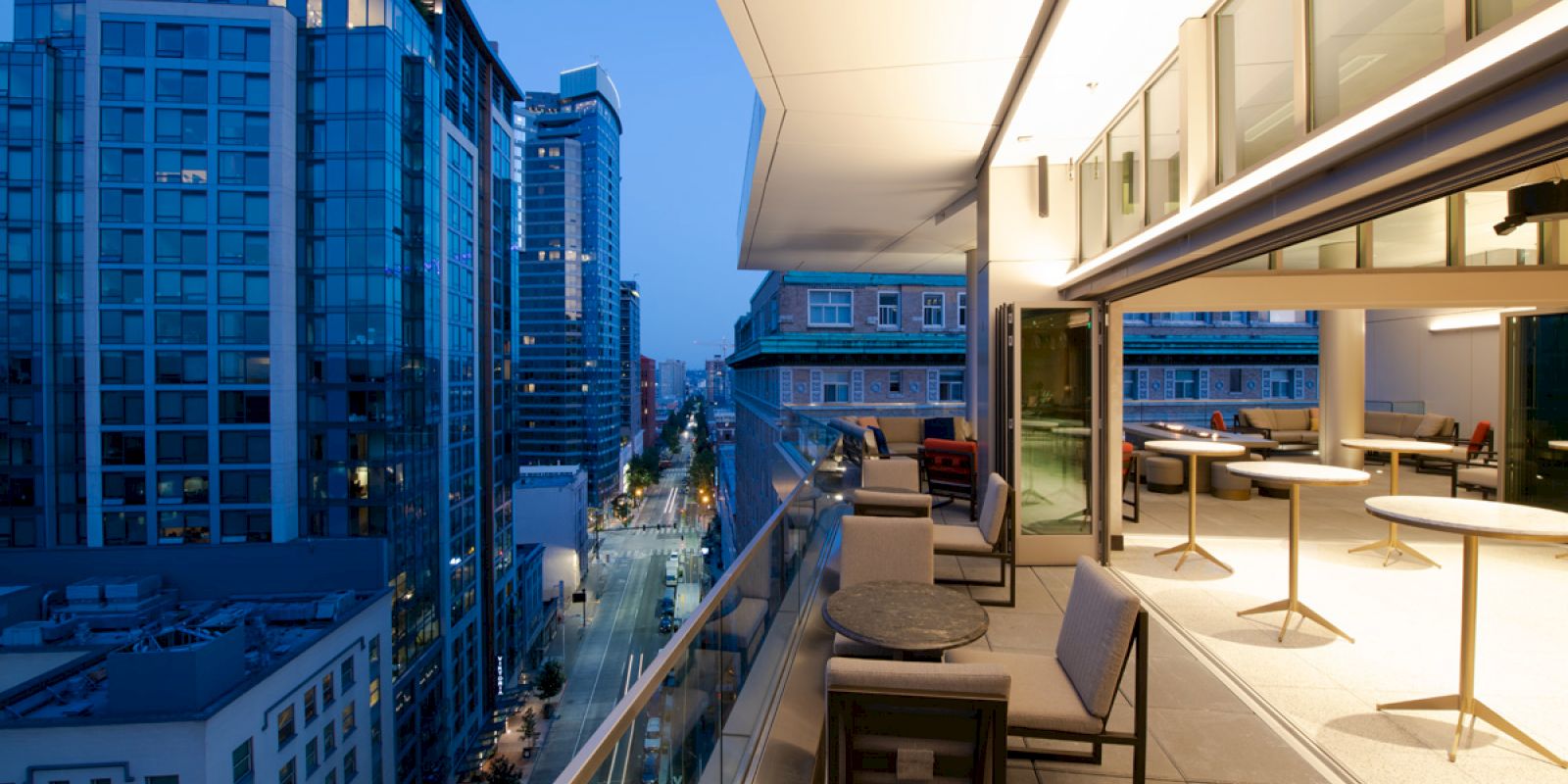 A modern cityscape with tall buildings, viewed from a spacious balcony with seating and tables, during the evening with city lights glowing.