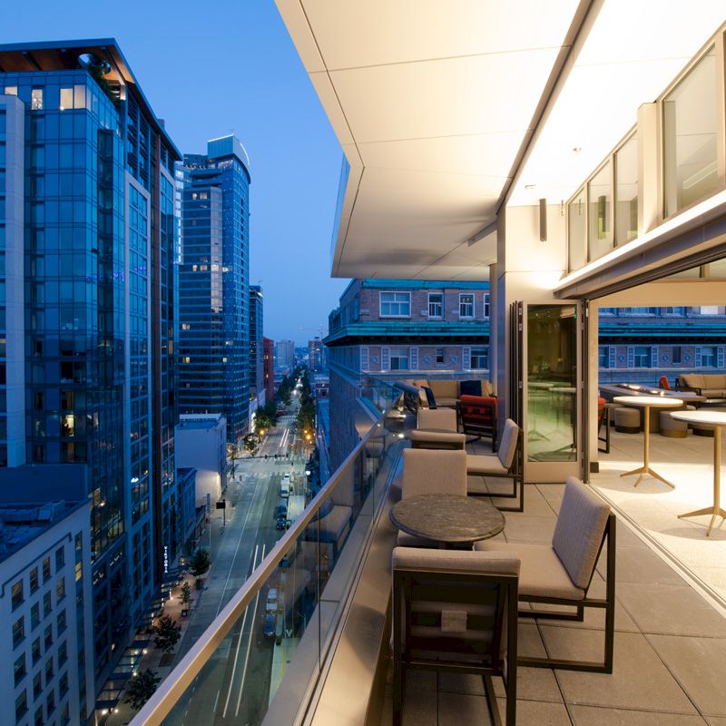 A modern balcony with seating and tables overlooks a city street lined with tall buildings, captured during the evening.