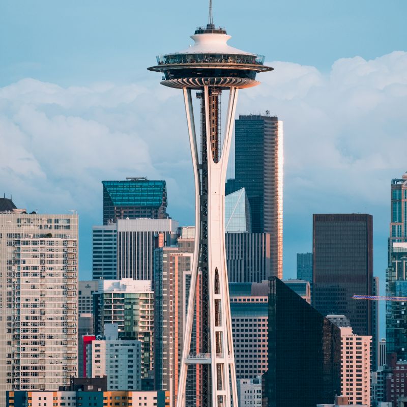 The image shows the Space Needle in Seattle, surrounded by modern city buildings under a partly cloudy sky, a significant urban landmark.