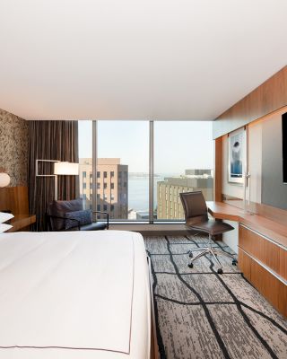 A modern hotel room featuring a large bed, wall-mounted TV, desk, chair, and a view of city buildings through large windows ending the sentence.