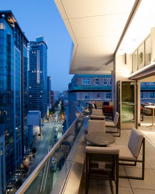 A modern cityscape at dusk with tall buildings, seen from a spacious balcony featuring tables and chairs, and a brightly lit interior space.