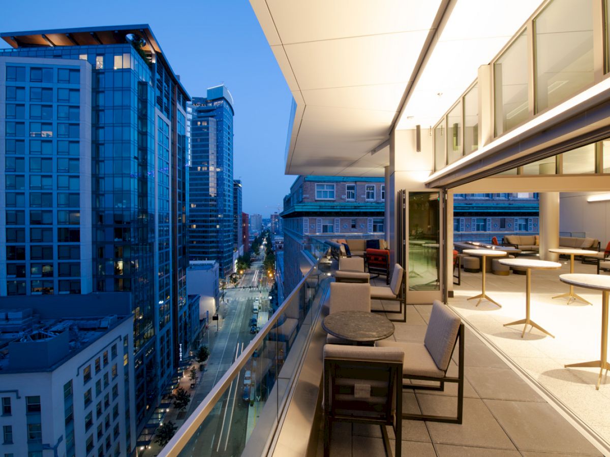 A modern cityscape at dusk with high-rise buildings and a furnished balcony including tables, chairs, and a view of a street below.