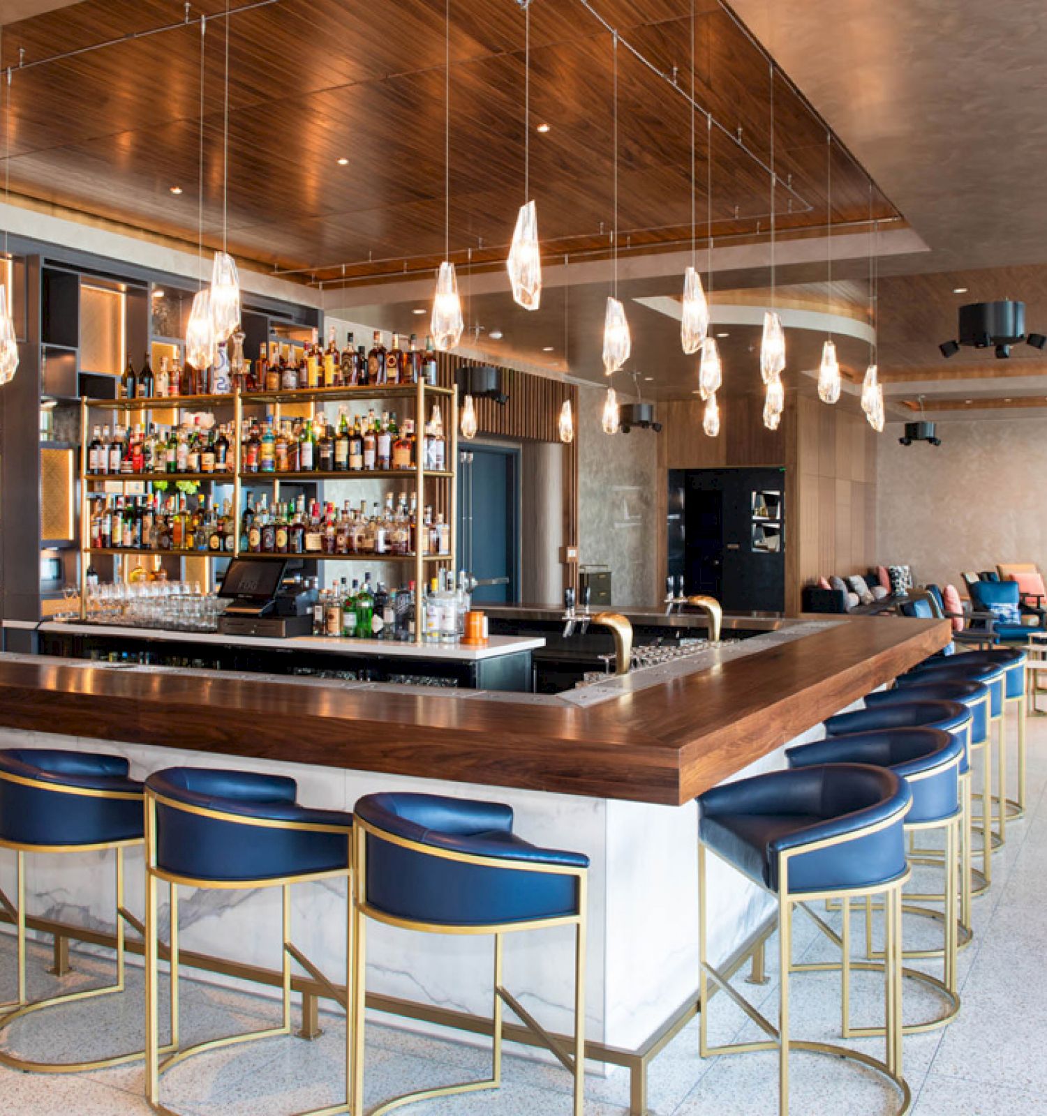The image shows a modern bar with a wooden counter, stylish stools, a wide selection of bottles, and elegant lighting. The seating area is visible.