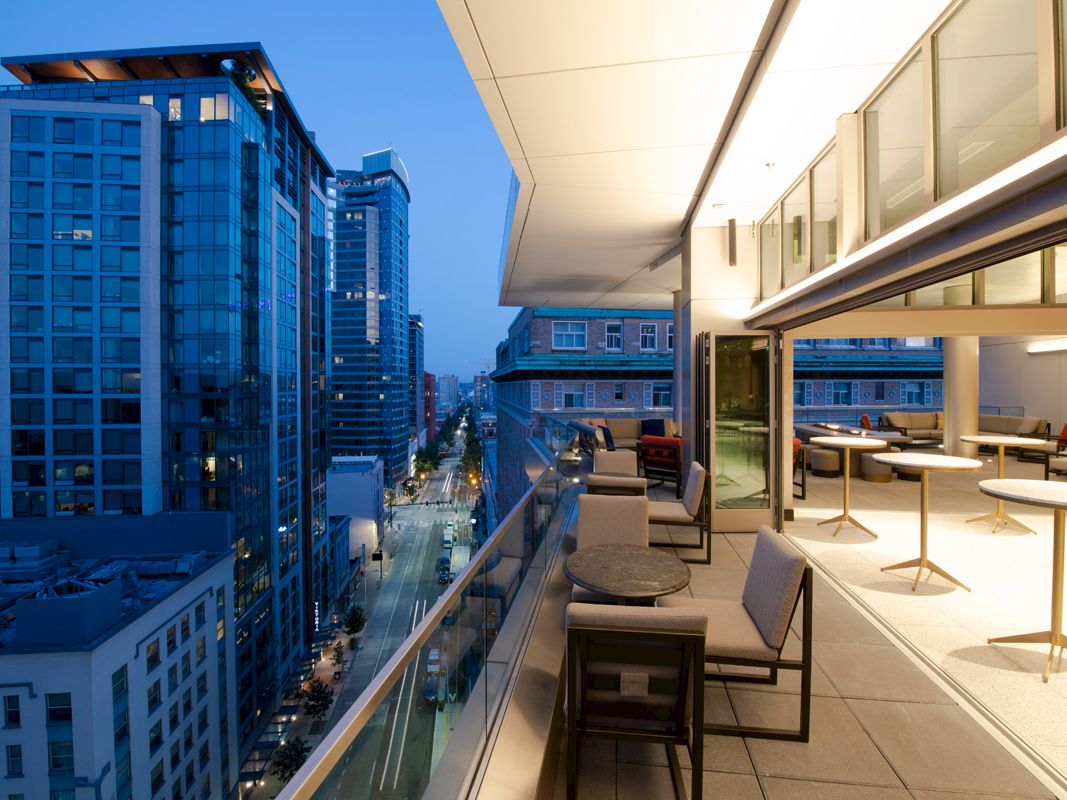 A modern outdoor terrace overlooking a city street at dusk. It has tables, chairs, and glass railing, with buildings visible in the background.