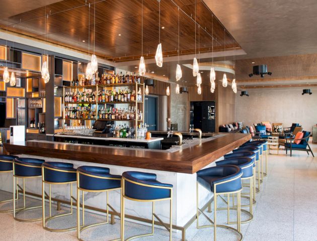 A modern bar with a wooden countertop, blue barstools, hanging pendant lights, a stocked bar, and seating area in the background.