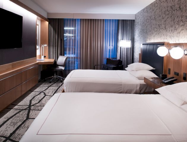 A modern hotel room with two double beds, a wall-mounted TV, a work desk with a chair, and large windows with curtains.