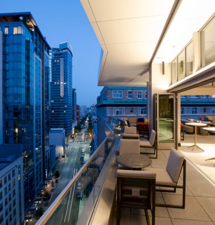 A modern balcony overlooking a cityscape at dusk, with buildings, streetlights, and a well-lit outdoor seating area featuring tables and chairs.