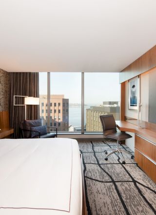 A modern hotel room features a king bed, large TV, desk, and chair with floor-to-ceiling windows offering a cityscape view and partial water view.
