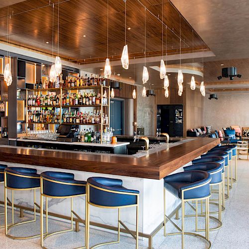 A modern bar setup with a wooden counter, blue chairs, and pendant lights hanging from the ceiling, with a well-stocked bar in the background.