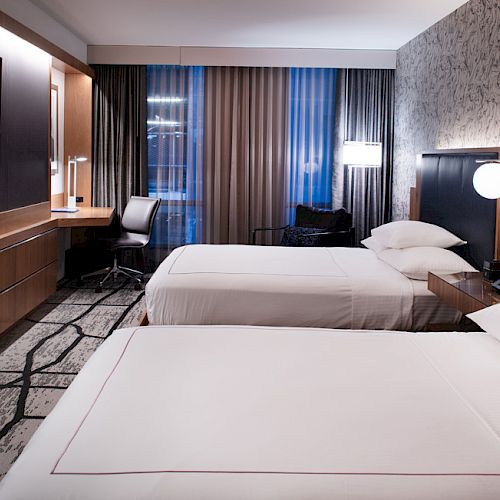 A modern hotel room with two neatly made beds, a flat-screen TV, a desk with a chair, and ambient lighting.