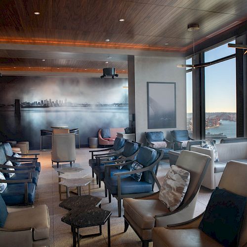 A modern lounge room with ample seating, large windows offering a scenic waterfront view, and a cityscape mural on the back wall.